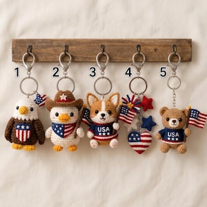 May include: A collection of five crochet keychains with patriotic themes. Each keychain features a different animal or object, including an eagle, duck, corgi, heart, and bear, all holding American flags. The keychains are attached to a wooden rack.