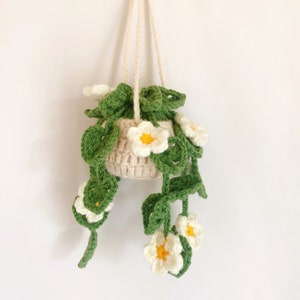May include: A crocheted hanging planter with green leaves and white flowers. The planter is made of beige yarn and has a hanging loop.