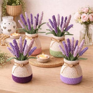 Puede incluir: Cuatro jarrones de ganchillo hechos a mano, cada uno con ramitas de lavanda. Los jarrones son blancos con fondos de colores y un acento de corazón de madera. Una pequeña vela se encuentra sobre una estera tejida, y un jarrón de rosas rosas está en el fondo.