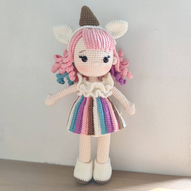 K&ouml;nnte beinhalten: Eine geh&auml;kelte Puppe mit rosa Haaren, einem gestreiften Kleid und einer braunen und wei&szlig;en Eiswaffel-M&uuml;tze.