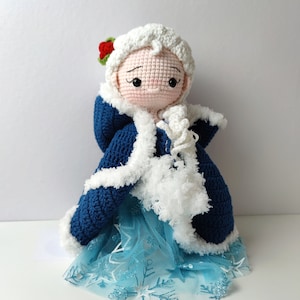 Pu&ograve; includere: Una bambola all'uncinetto che indossa un mantello blu con cappuccio e bordatura in pelliccia bianca e una gonna di tulle blu con motivi a fiocco di neve. La bambola ha i capelli bianchi all'uncinetto e un viso rosa con occhi dipinti e un fiore rosso sulla testa.