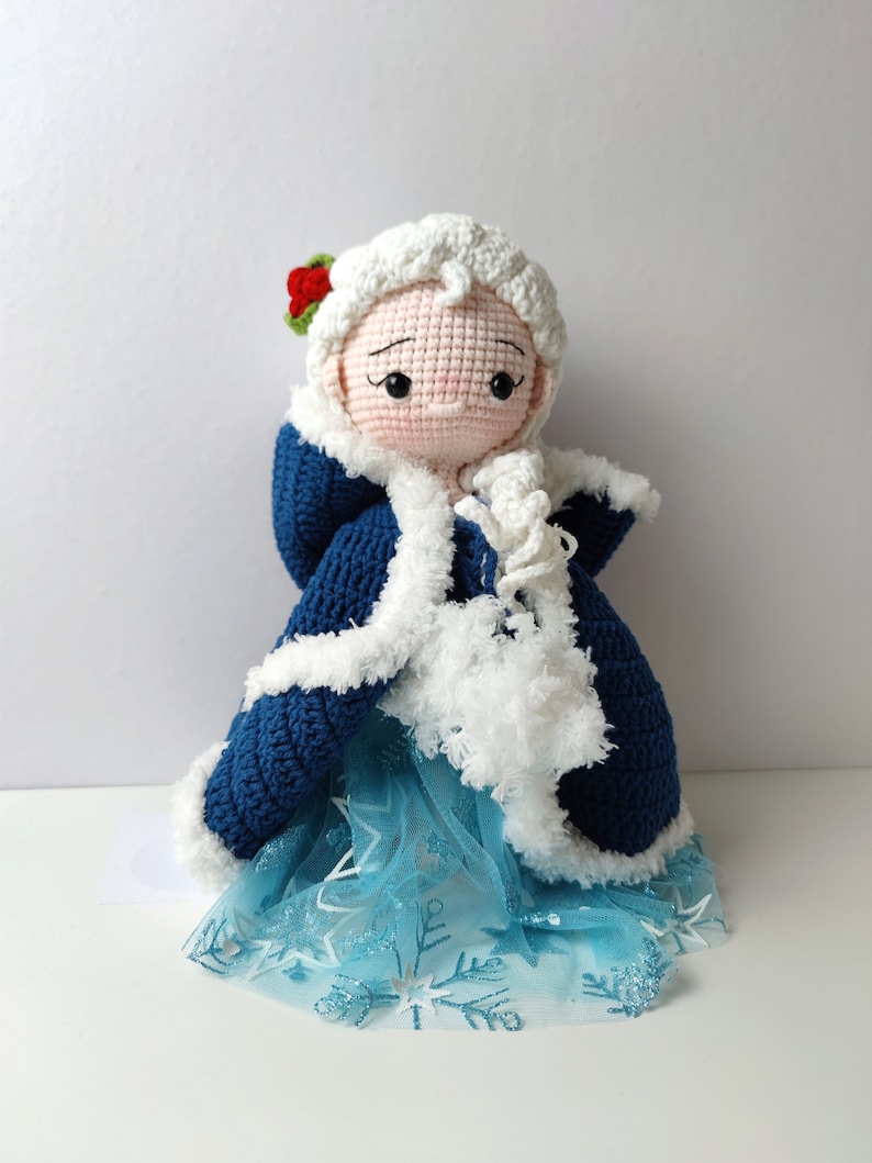Pu&ograve; includere: Una bambola all'uncinetto che indossa un mantello blu con cappuccio e bordatura in pelliccia bianca e una gonna di tulle blu con motivi a fiocco di neve. La bambola ha i capelli bianchi all'uncinetto e un viso rosa con occhi dipinti e un fiore rosso sulla testa.