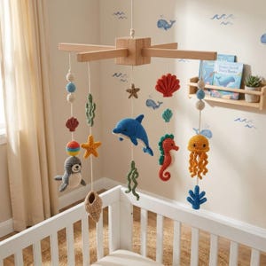 May include: A wooden nursery mobile featuring crocheted sea creatures including a blue dolphin, orange seahorse, yellow jellyfish, gray seal, and various shells, starfish, and seaweed shapes suspended from a central wooden cross frame.
