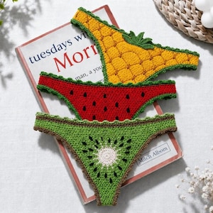 May include: Three crochet bikini bottoms designed to resemble fruit. The top piece is yellow with a pineapple texture and green leaves. The middle piece is red with black seeds like a watermelon. The bottom piece is green with a white center.