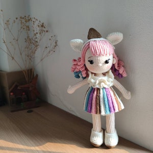 K&ouml;nnte beinhalten: Eine geh&auml;kelte Puppe mit rosa Haaren, wei&szlig;en Ohren und einem bunten, gestreiften Rock. Die Puppe tr&auml;gt ein wei&szlig;es, geh&auml;keltes Oberteil mit einem R&uuml;schenkragen.