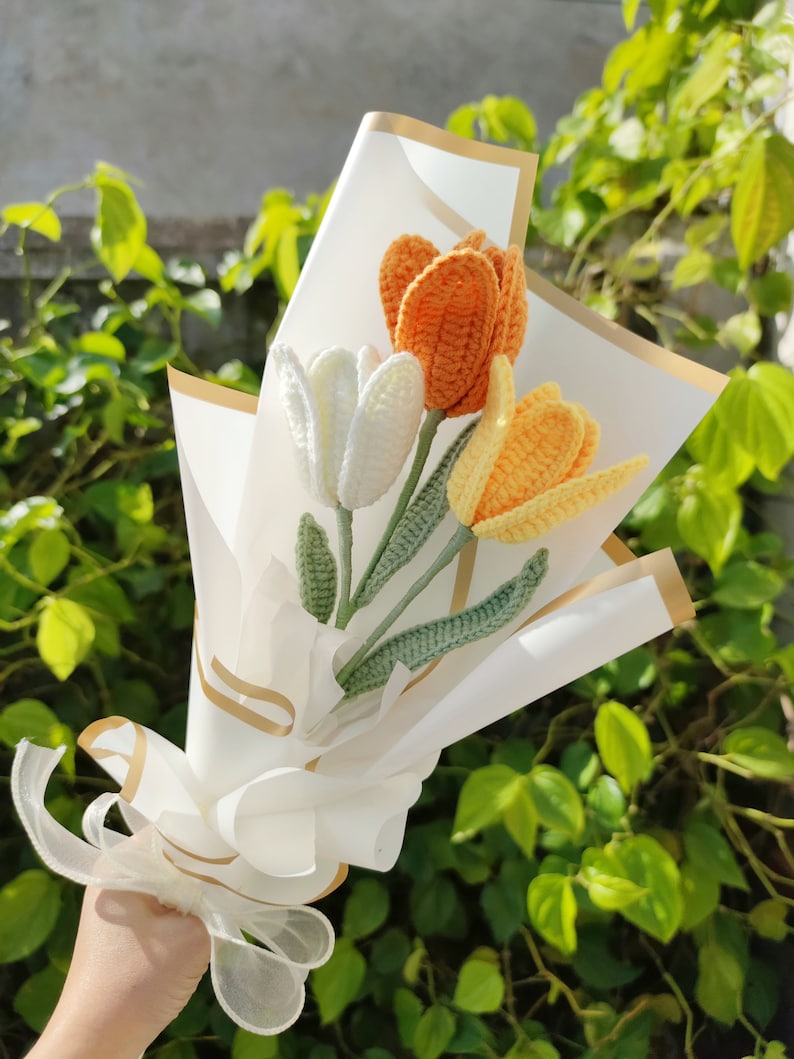 May include: A bouquet of crocheted tulips in white, orange, and yellow, wrapped in white paper with a gold ribbon.