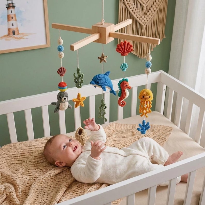 May include: A wooden baby mobile featuring crocheted sea creatures including a seal, dolphin, seahorse, jellyfish, starfish, and seaweed, suspended from a cross-shaped frame with colorful yarn balls and shells hanging above a crib.