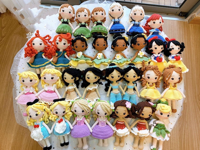 Pu&ograve; includere: Una collezione di bambole principesse Disney all'uncinetto. Le bambole indossano tutte i loro abiti caratteristici e hanno tratti del viso dettagliati. Le bambole sono disposte in cerchio su una superficie bianca.