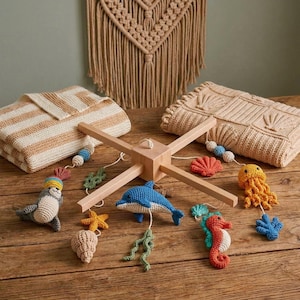 May include: A wooden baby mobile featuring crocheted sea creatures including a blue dolphin, orange seahorse, yellow jellyfish, and a seal, alongside crocheted shells, starfish, and seaweed, suspended from a central light wood cross frame.