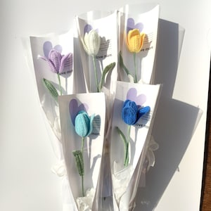 May include: A bouquet of five crocheted tulips in different colors, including purple, white, yellow, blue, and light blue. Each tulip is wrapped in a clear plastic sleeve with a white card that says "love more".