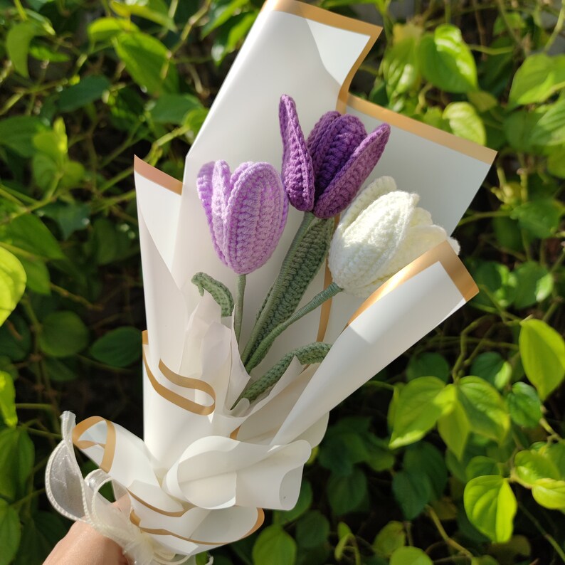May include: A bouquet of three crocheted flowers in a white paper wrapper with a gold ribbon. The flowers are purple and white.