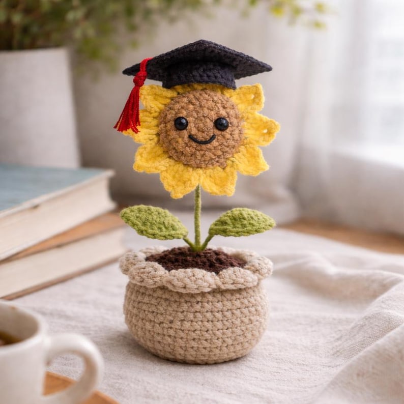 Puede incluir: Un girasol de ganchillo con un birrete de graduaci&oacute;n con una borla roja. La flor amarilla tiene una cara sonriente y hojas verdes. Est&aacute; plantado en una maceta de ganchillo beige llena de tierra marr&oacute;n.