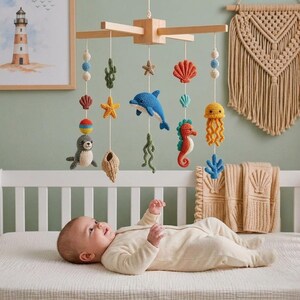 May include: A wooden baby mobile featuring crocheted sea creatures including a dolphin, seahorse, seal, jellyfish, starfish, and seashells suspended by white strings. The mobile includes colorful yarn beads and is hung above a baby lying on a bed.