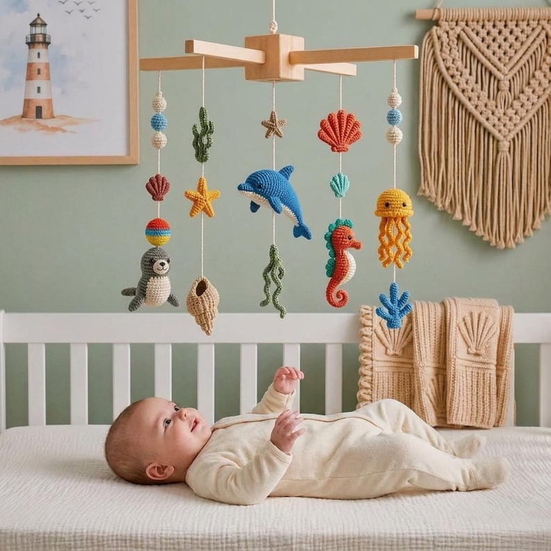 May include: A wooden baby mobile featuring crocheted sea creatures including a dolphin, seahorse, seal, jellyfish, starfish, and seashells suspended by white strings. The mobile includes colorful yarn beads and is hung above a baby lying on a bed.