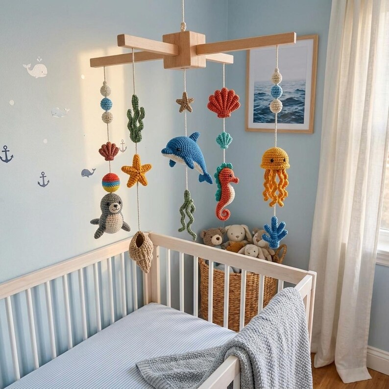 May include: A wooden baby mobile featuring crocheted sea creatures including a gray seal, blue dolphin, orange seahorse, yellow jellyfish, starfish, shells, and seaweed hanging from white strings attached to a cross-shaped wooden frame.