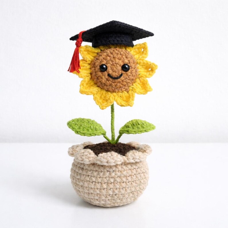 Puede incluir: Un girasol tejido a ganchillo con un birrete de graduaci&oacute;n con una borla roja. La flor amarilla tiene una cara sonriente y est&aacute; plantada en una maceta beige con hojas verdes. Un encantador regalo hecho a mano para un graduado.