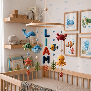May include: A wooden baby mobile featuring crocheted sea creatures, such as a whale, turtle, crab, and octopus, suspended above a cot. The mobile spells "LIAM" with blue letters. Framed ocean-themed artwork decorates the wall.