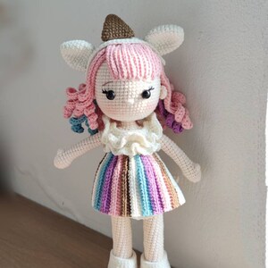 K&ouml;nnte beinhalten: Eine geh&auml;kelte Puppe mit rosa Haaren, einem gestreiften Rock und wei&szlig;en Schuhen. Die Puppe tr&auml;gt einen braunen und wei&szlig;en Hut mit Hasenohren.