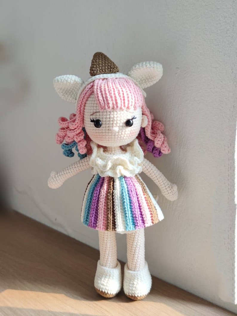 K&ouml;nnte beinhalten: Eine geh&auml;kelte Puppe mit rosa Haaren, einem gestreiften Rock und wei&szlig;en Schuhen. Die Puppe tr&auml;gt einen braunen und wei&szlig;en Hut mit Hasenohren.
