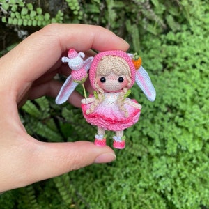 May include: A small crocheted doll wearing a pink dress and a pink bunny hat. The doll is holding a white bunny with a pink flower.