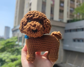 May include: A brown crocheted poodle toy with a blue bell around its neck. The toy is being held in a person's hand.