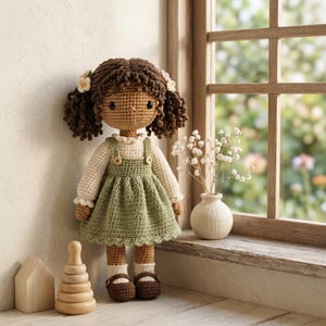 Könnte beinhalten: Eine handgefertigte Häkelpuppe mit dunkelbraunem, lockigem Haar, das mit kleinen weißen Blumen verziert ist. Die Puppe trägt ein cremefarbenes Langarmshirt unter einem salbeigrünen Trägerkleid mit zwei Knöpfen sowie weiße Socken und braune Schuhe.