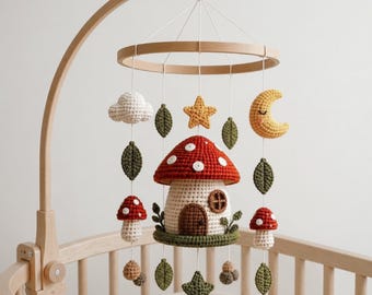 May include: A crochet baby mobile featuring a central mushroom house, surrounded by hanging elements including a white cloud, a yellow crescent moon, green leaves, small red mushrooms, and stars, all suspended from a circular wooden frame.