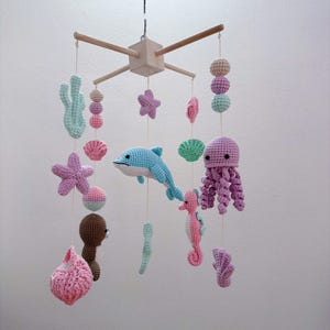 May include: A hanging nursery mobile featuring crocheted sea creatures including a blue dolphin, a pink seahorse, a purple octopus, starfish, shells, and coral in pastel shades. The figures are suspended from a light wooden cross frame.