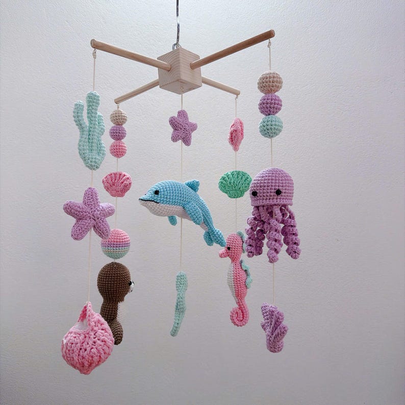 May include: A hanging nursery mobile featuring crocheted sea creatures including a blue dolphin, a pink seahorse, a purple octopus, starfish, shells, and coral in pastel shades. The figures are suspended from a light wooden cross frame.