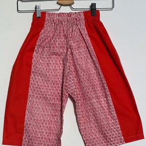 May include: Red and pink patterned shorts hanging on a metal hanger. The shorts have solid red panels on the sides and a pink and white geometric pattern in the center. The shorts are made of a lightweight fabric.