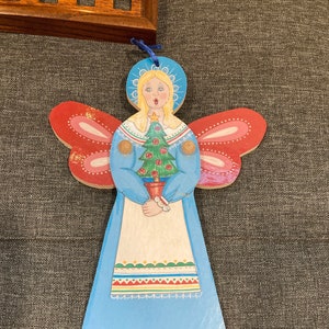 May include: Wooden Christmas angel ornament with a blue dress, red wings, and a blonde halo. The angel holds a green Christmas tree in a pot. The ornament has a blue string for hanging and is decorated with floral and gold accents.