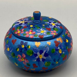 May include: A blue, hand-painted wooden trinket box with a lid. The box is adorned with colorful floral and heart designs in pink, red, yellow, and white. The lid has a small, rounded knob.