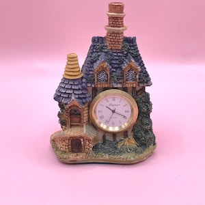 May include: A decorative table clock shaped like a cottage with a stone-textured facade, a tall brick chimney, and a blue shingled roof. The round clock face features a gold-tone bezel, black hands, and the word Elegance printed in black.