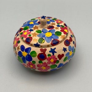 May include: A round, beige ceramic jar with a lid, adorned with hand-painted flowers in blue, pink, and yellow. The lid features a small knob. The jar's exterior is covered in colorful floral designs, creating a cheerful, decorative piece.