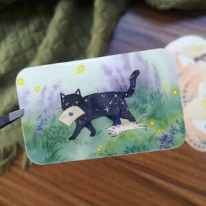 Peut inclure: Autocollant rectangulaire représentant un chat noir avec un motif étoilé portant une lettre et une petite souris courant à proximité. Le sticker a un design fantaisiste avec un fond violet et vert, tenu par une pince.