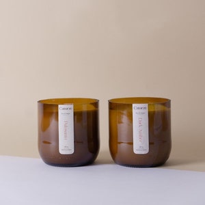 Puede incluir: Dos tarros de velas de vidrio ámbar con etiquetas blancas. La vela de la izquierda está etiquetada como "Palo Santo" y la vela de la derecha está etiquetada como "Dark Amber". Ambas velas son de soja y veganas.