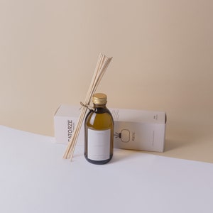 Könnte beinhalten: Eine braune Glasflasche mit goldenem Deckel und einem Etikett mit der Aufschrift "poetic" und "atelier". Die Flasche ist mit einer klaren Flüssigkeit gefüllt und hat Holzstäbchen, die aus ihr herausragen. Die Flasche steht auf einer weißen Oberfläche neben einer weißen Schachtel mit der Aufschrift "ATORZE".