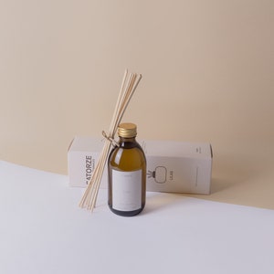 Op de afbeelding: Een bruine glazen fles met een gouden dop en een wit etiket met de tekst "Fig" is gevuld met een heldere vloeistof. De fles is omgeven door houten stokjes en een witte doos met de tekst "LEIAS" erop.