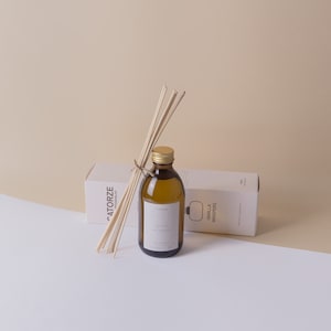 Op de afbeelding: Een bruine glazen fles met een gouden dop en een wit etiket met de tekst "Vanilla Whispers" is omgeven door houten stokjes. De fles staat op een wit oppervlak met een witte doos waarop "Catorze Fragrances" en "Vanilla Whispers" staat.