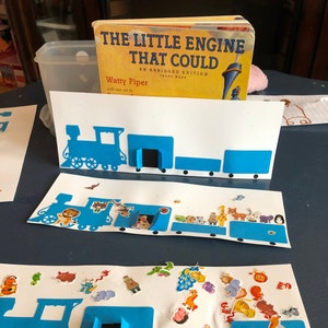 Könnte beinhalten: Eine Bastelarbeit auf weißem Papier mit einer blauen Lokomotive und mehreren Waggons. Der Zug ist mit bunten Tieraufklebern verziert. Das Buch "Die kleine Lokomotive, die konnte" ist im Hintergrund zu sehen.
