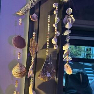 Puede incluir: Un móvil de conchas marinas hecho a mano. El móvil presenta varias conchas marinas y cuentas suspendidas de una rama natural. Las conchas son en tonos de marrón, beige y blanco, con cuentas doradas y nacaradas. El móvil está diseñado para uso en interiores.