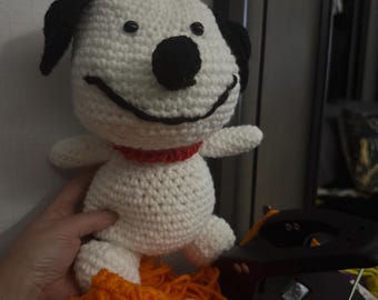 May include: A handmade white and black crochet dog with a red collar. The dog has a smiling face and black button eyes. The dog is part of a collection of crochet items, including an orange hat and pink yarn.