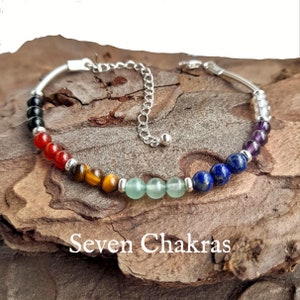 May include: A silver bracelet with seven chakra stones. The stones are black, red, brown, green, blue, purple, and white. The bracelet has a silver clasp and is on a wooden surface. Seven Chakras.