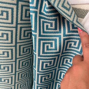 May include: Close-up of a teal and off-white patterned fabric. The fabric features a repeating geometric design of interlocking squares. The pattern is a vibrant teal against a neutral off-white background. The fabric is folded, revealing the pattern's depth.