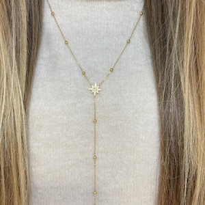 Peut inclure: Un collier doré avec un pendentif en forme d'étoile et une chaîne délicate. Le collier comporte de petites perles rondes le long de la chaîne et une goutte plus longue avec des perles supplémentaires. Le bijou est porté sur un pull crème.