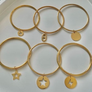 May include: Gold bangles with charm pendants in star, lightning bolt, and circular designs. The bracelets are presented on a white tray, highlighting their simple, elegant design. These accessories are ideal for adding a touch of sparkle to any outfit.