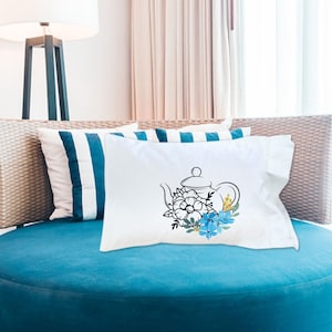 May include: White pillow with a black line drawing of a teapot with blue and yellow flowers. The pillow is on a blue couch.