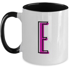 Könnte beinhalten: Weiße Keramik-Kaffeetasse mit schwarzem Rand und Henkel. Die Tasse zeigt einen pinkfarbenen Buchstaben "E" mit schwarzen Umrissen.