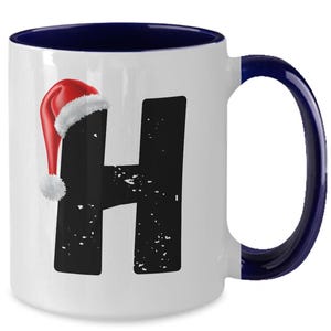 May include: White ceramic mug with a navy blue interior and handle. The letter "H" is printed in black with a distressed texture, and a Santa hat is on the top left. A festive holiday mug.