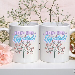 Puede incluir: Dos tazas de cerámica blanca con un diseño rosa, verde y azul. Las tazas tienen el texto "Let's get Egg-cited!"